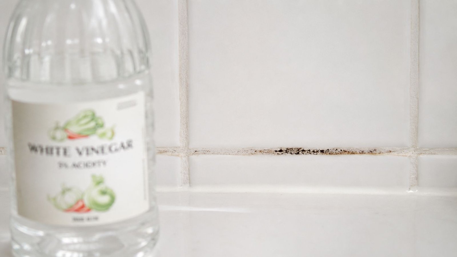 A bottle of white vinegar sitting on a kitchen counter near grout with visible black mold spots.